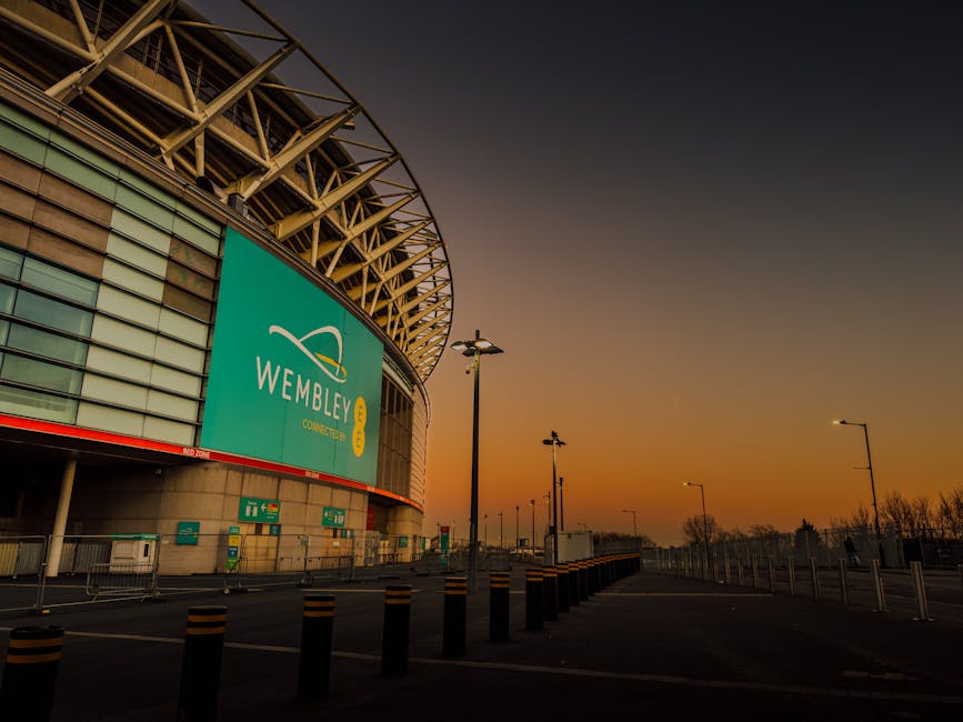 Inside Wembley Stadium, the view shows a large football pitch with lush green grass, surrounded by multiple tiers of bright red seating. In the foreground, there are several large cardboard boxes and 