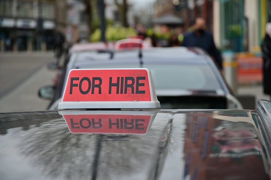 A close-up view of a black van with a shiny, reflective surface displaying a prominent red and white sign reading 'FOR HIRE' mounted on the roof, indicating its availability for house removals or movi